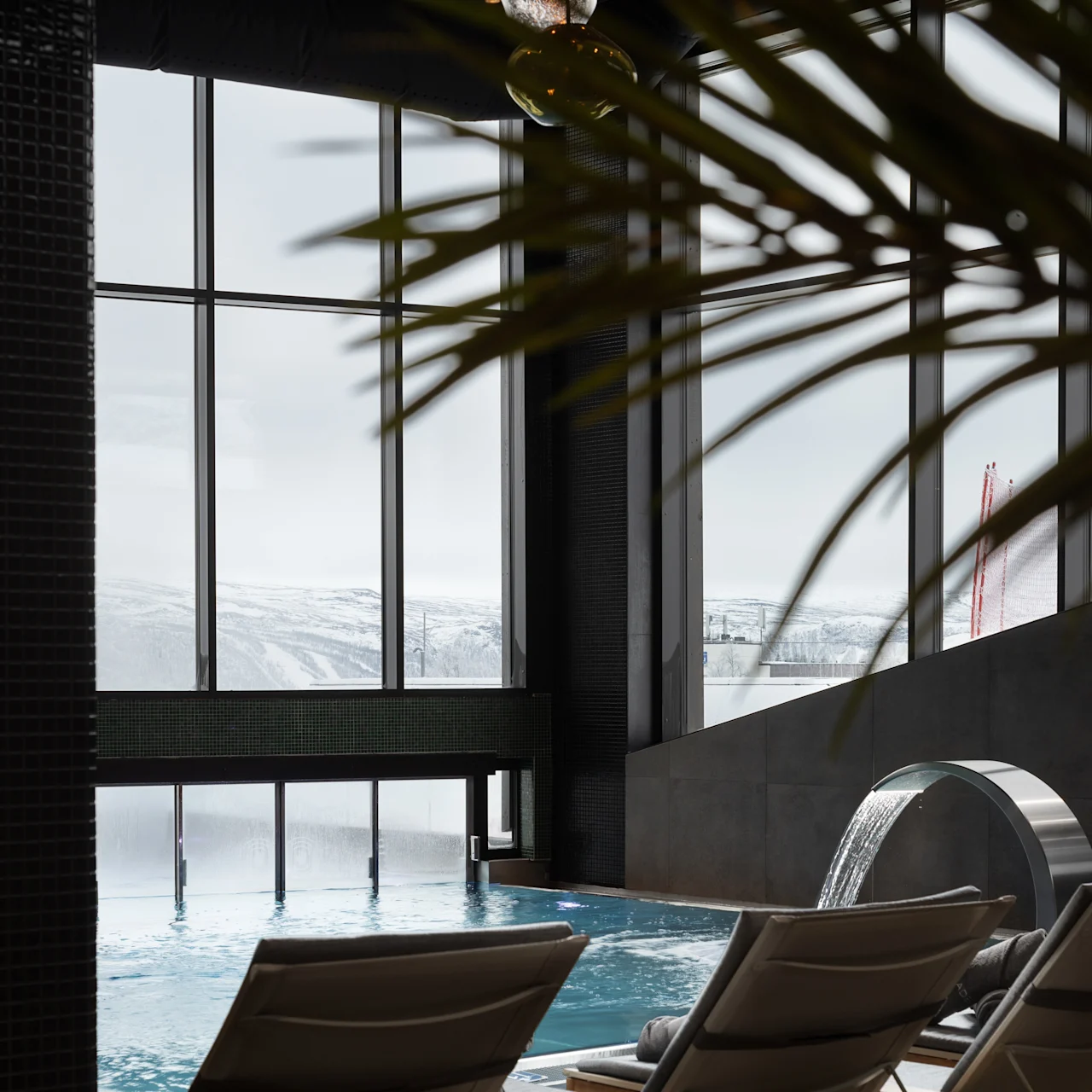 Liggestole vender mod en indendørs pool med rindende vand, mod store vinduer, der viser et snedækket landskab udenfor. Overhead giver elegante pendellamper et varmt skær.