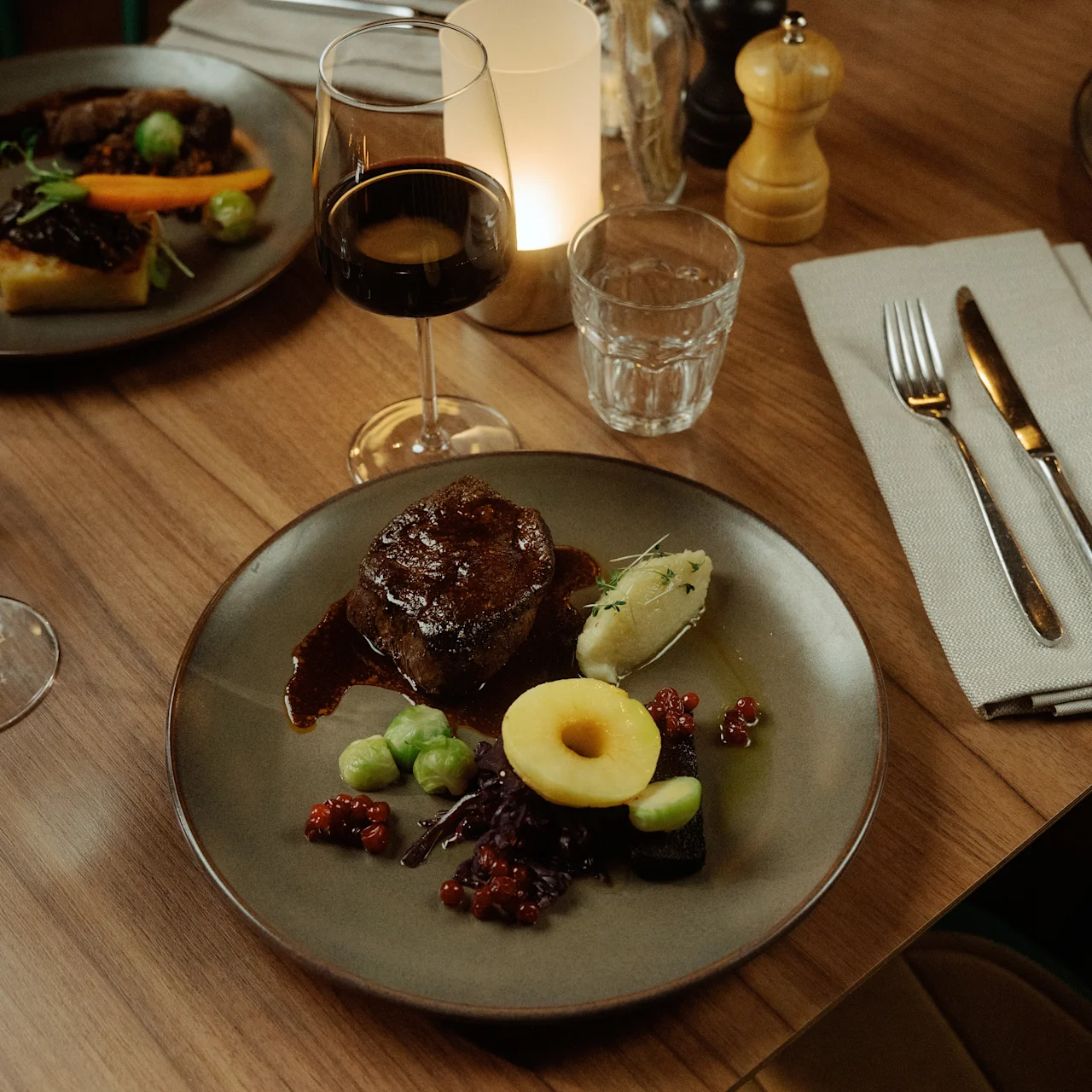 Tallrik med biff, sås, potatis och garnering; bredvid vin-, vattenglas, bestick och ljus på träbord i restaurangmiljö för en elegant middag.