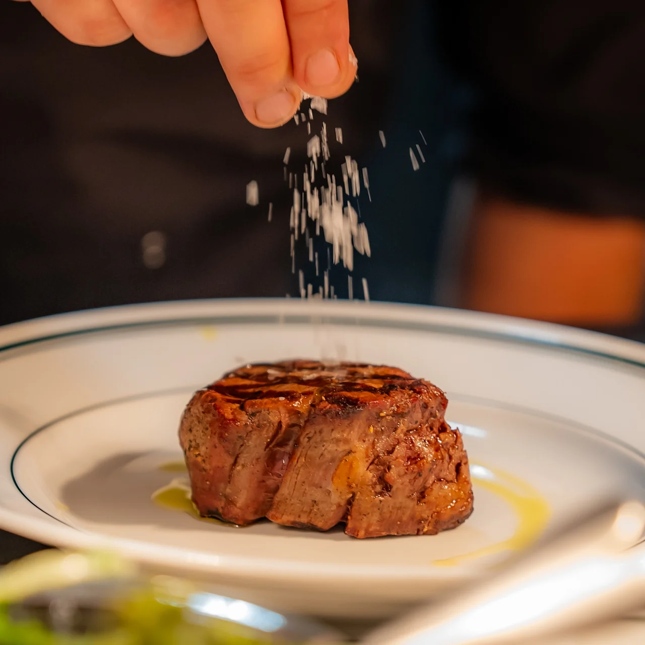En hand strör salt över en grillad biff på en vit tallrik med gröna linjer i ett svagt upplyst kök.