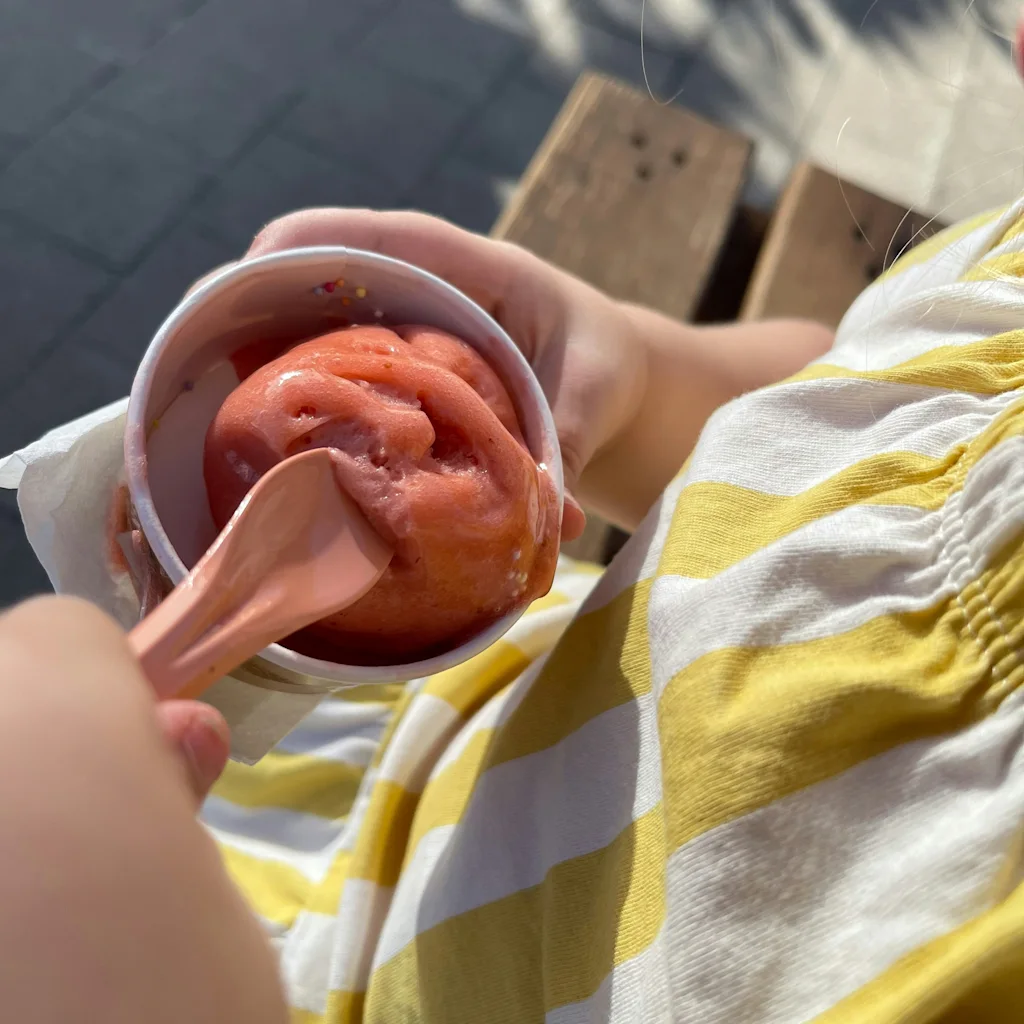 En person i en randig tröja håller en kopp med rosa sorbet eller glass med en sked, njuter av en godsak utomhus.