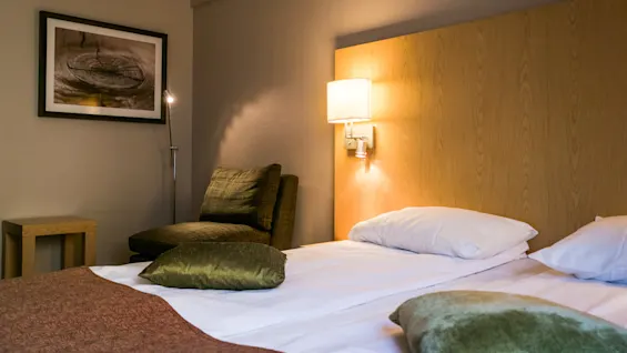 Bed with a wooden headboard, adorned with pillows and blankets, situated in a warmly-lit, cozy bedroom with an armchair and wall art. A lamp shines softly overhead.