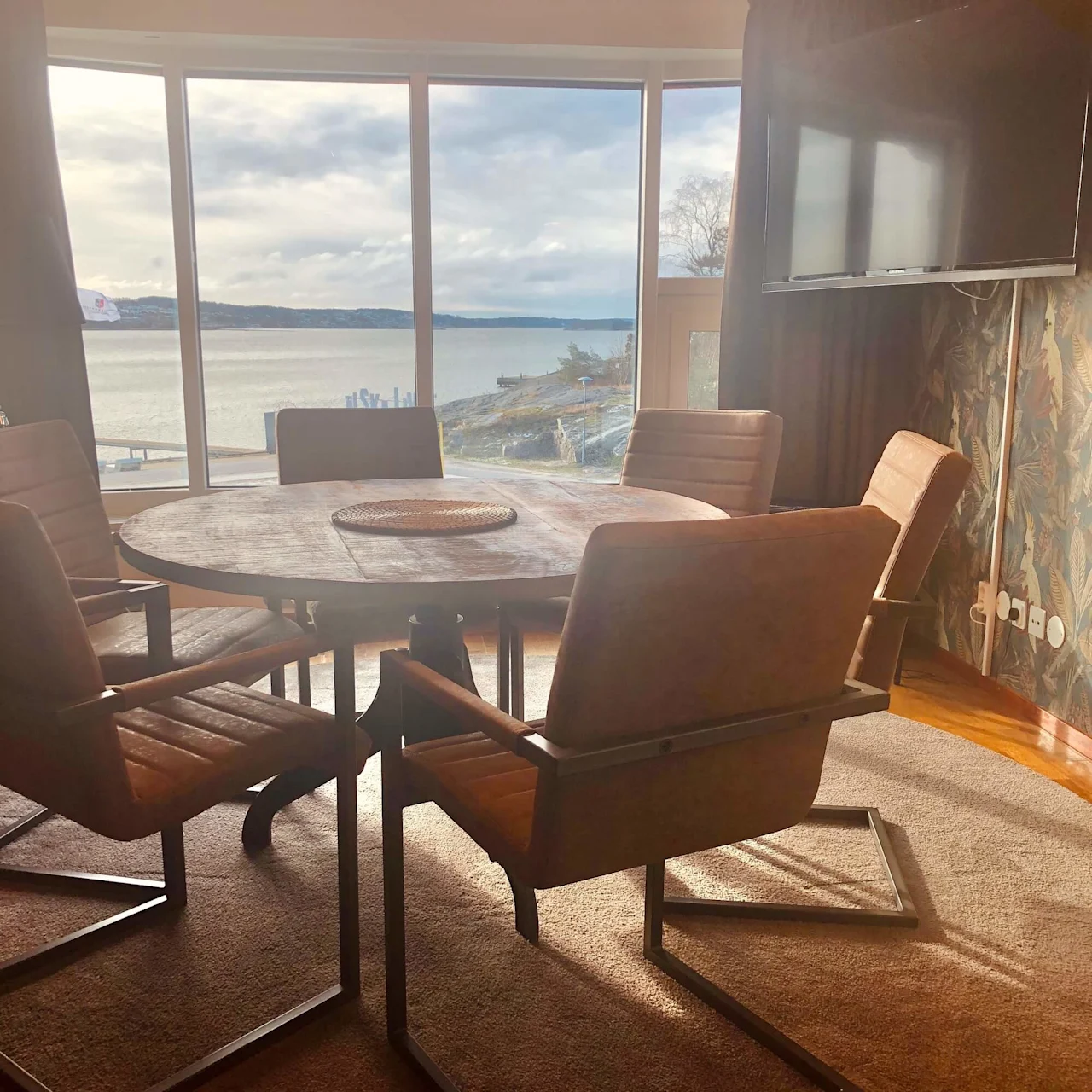 A round wooden table surrounded by five cushioned chairs sits in a sunlit room with a large window. Outside, a scenic view of a lake and cloudy sky.