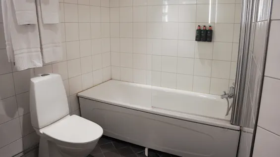 A bathroom with a toilet and bathtub/shower with glass screen, white towels hanging, and tiled walls.
