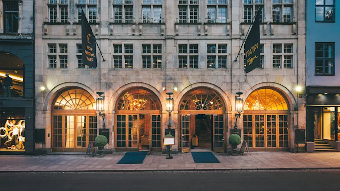 A grand building facade features three arched wooden entrances, illuminated by amber lights. Two flags hang vertically, reading “HOTEL CRISTINA TEATRO” and “TEATRO”. Mannequins display clothes in the left shop window.