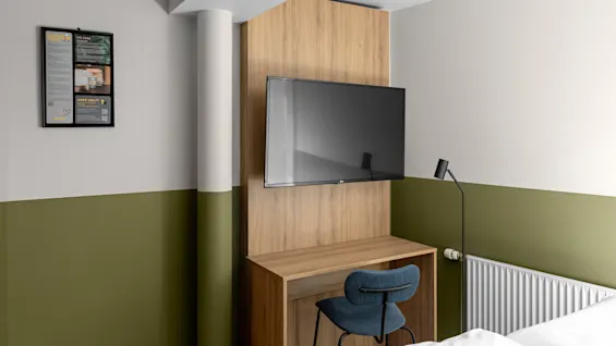 TV monterad på träpanel ovanför skrivbord med grön stol i rum med gröna och vita väggar. Inramat meddelande på väggen lyder: "Vi bryr oss. Behöver hjälp?" Golvlampa står bredvid en radiator.