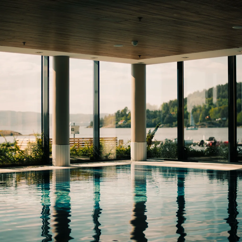 En inomhuspool med stora fönster som erbjuder en naturskön utsikt över en sjö omgiven av frodiga gröna kullar under en ljus himmel.