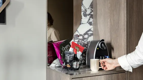 A hand holding a mug next to a coffee machine on a wooden countertop. Nearby, a basket contains snacks like bags labeled "CHIPS" and "NUTS."