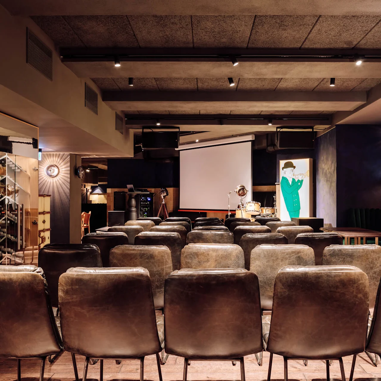 Leather chairs arranged facing a projector screen in a modern interior space with dim lighting and a vintage decor theme. A colorful illustration of a musician is visible on the right.