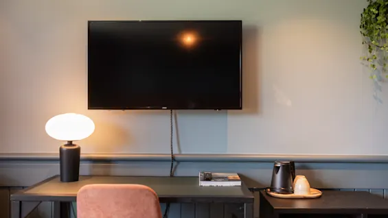 TV er monteret på en væg over et skrivebord med en lampe, bog, krus og kedel. En stol er skubbet ind under skrivebordet og en plante hænger i nærheden.