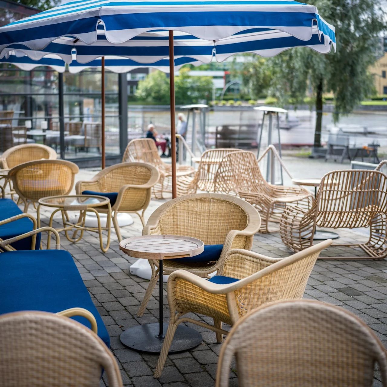 Korgstolar och bord placerade under ett randigt blått och vitt parasoll på en uteplats vid floden. Glasväggar omger en del av området med träd och byggnader i närheten.