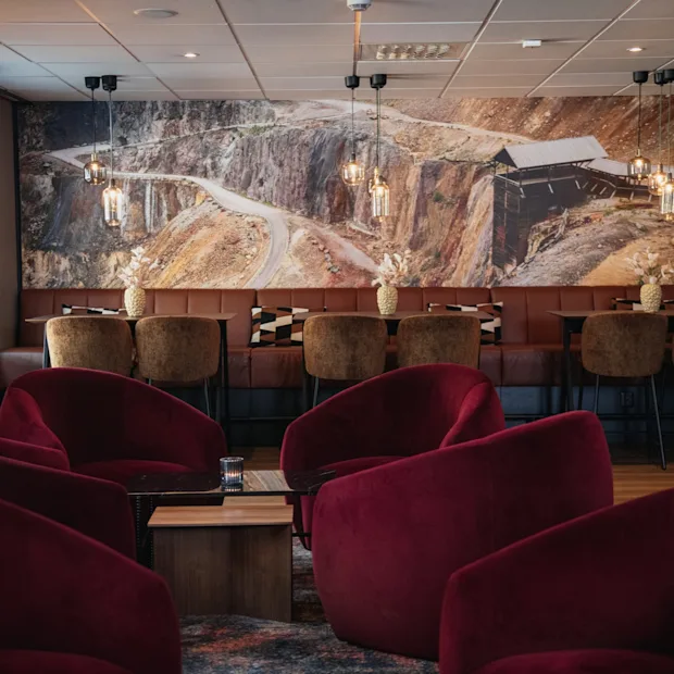 Et komfortabelt salongområde med myke røde lenestoler og benkeseter, med et stort veggmaleri av et ulendt landskap.