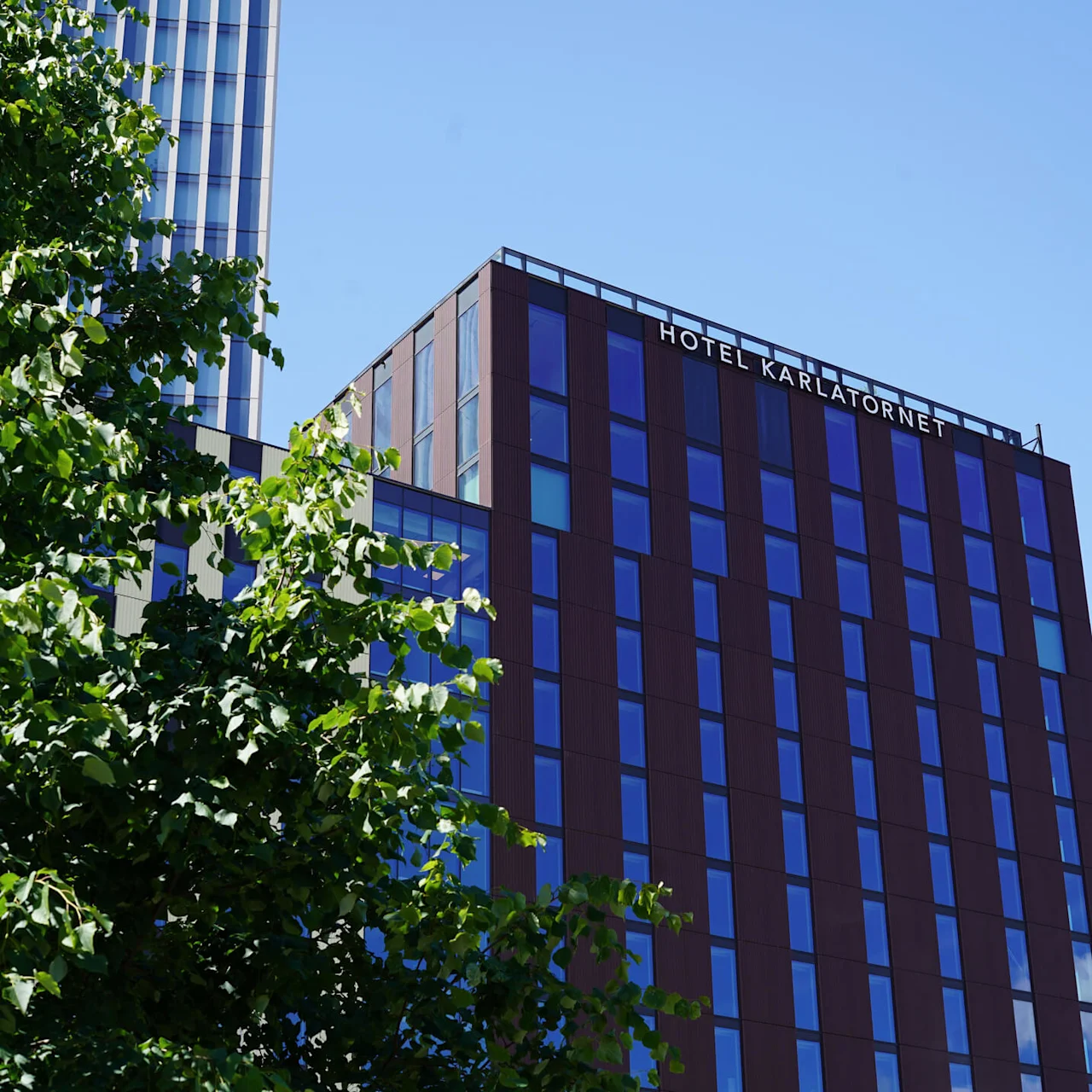En hög byggnad med texten "HOTEL KARLATORNET" står mot en klarblå himmel, omgiven av gröna lövträd.