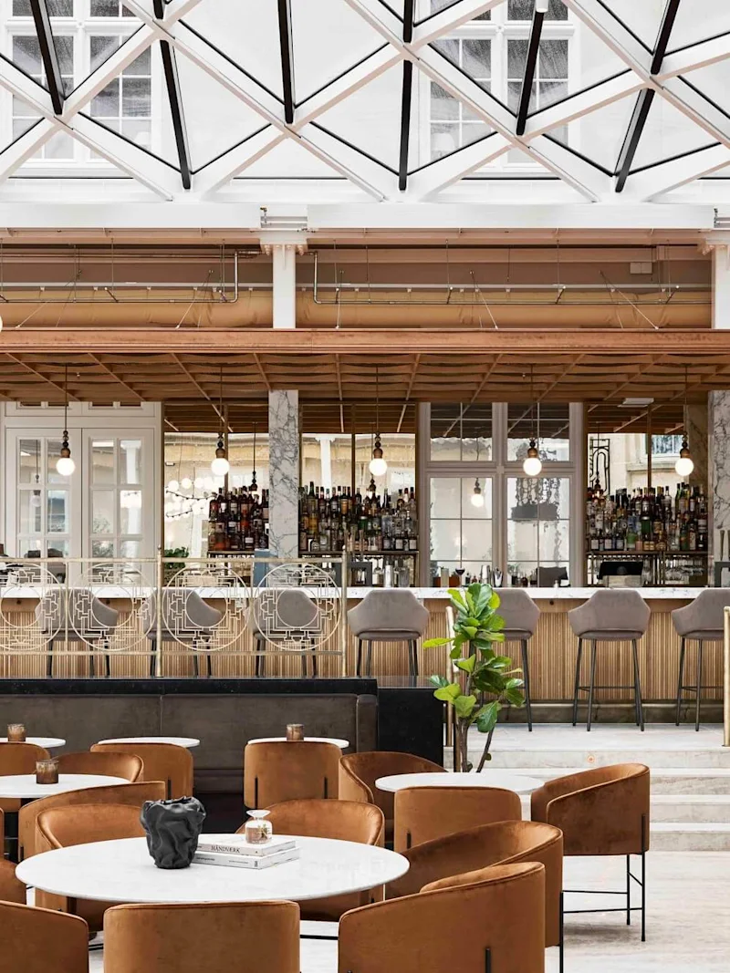 Bar med höga stolar under ett modernt glastak, omgiven av bruna loungestolar och runda bord i en stilfull, väl upplyst inomhusmiljö.