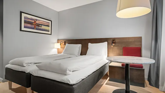 En pænt redt op tvillingeseng er placeret mod et træhovedgærde, ledsaget af et lille rundt bord og stol. En stor lampe hænger over, med et indrammet kunstværk på en lys væg.