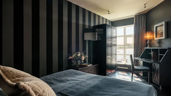 Et stilfuldt hotelværelse med en komfortabel seng, et mørkt træskrivebord ved et lyst vindue med udsigt over vandet og stribede vægge.