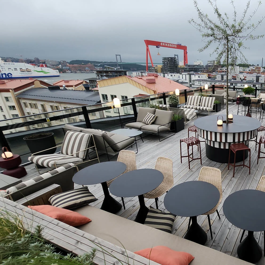 A rooftop terrace with modern furniture, tables, and seating areas overlooks a cityscape with a red crane labeled "ERIKSBERG" and a departing ship. Overcast sky creates a cozy atmosphere.