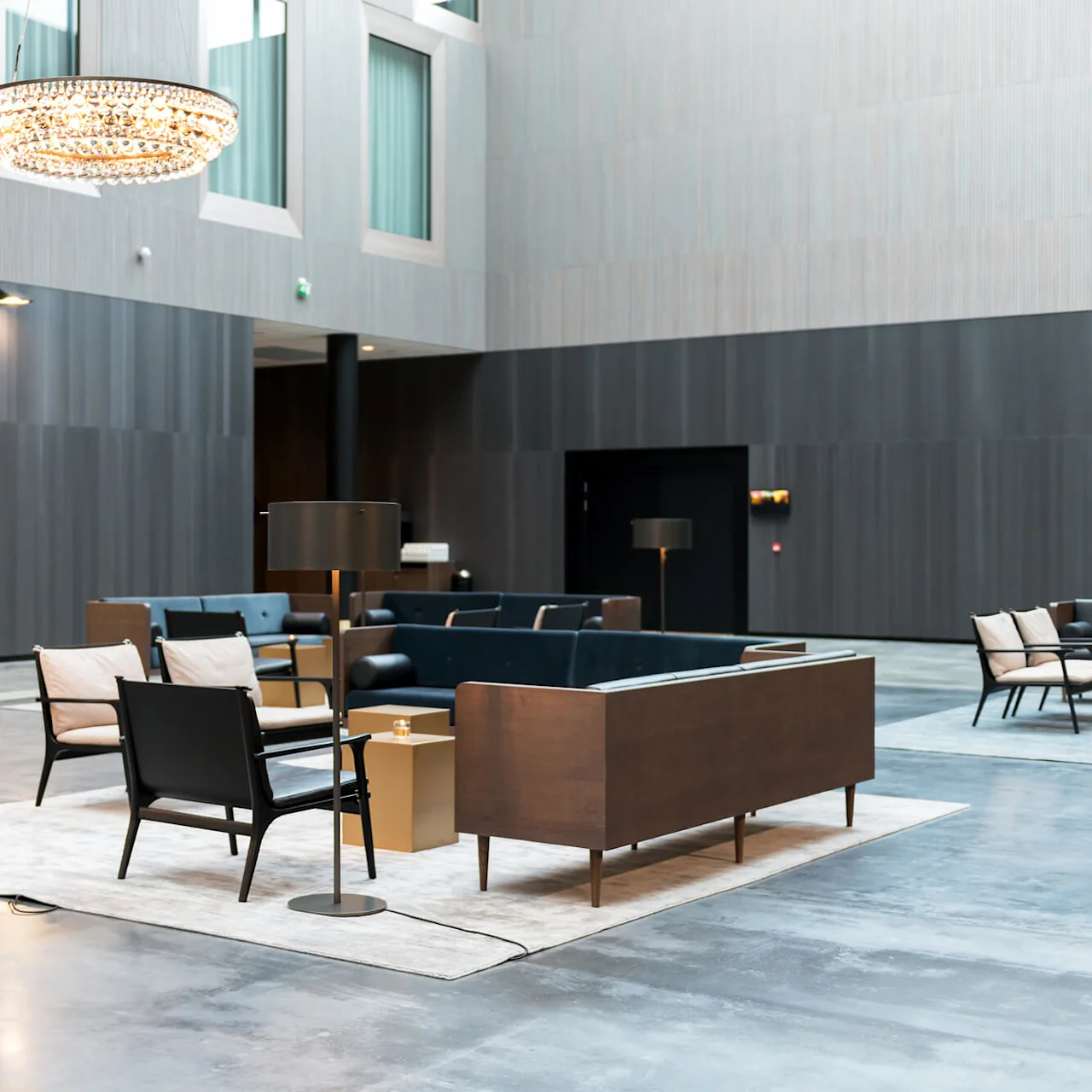 En sittegruppe med moderne møbler plassert på tepper, omgitt av høye mørke vegger, og opplyst av en stor, elegant lysekrone over.
