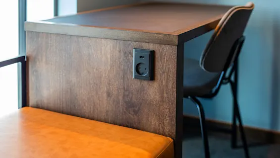 Strømuttak innebygd i en trebordsside. En USB-port og stikkontakt er synlig. Konteksten inkluderer en polstret brun benk og grå stol i et rom.