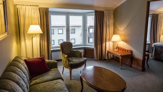 En stue har en vintage sofa og lenestol sentrert rundt et trebord. Varm belysning fra lamper forsterker den koselige atmosfæren, mens store vinduer har utsikt over nærliggende bygninger.