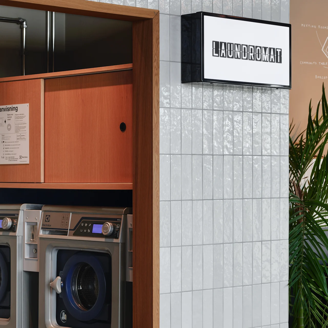 A laundromat with two washing machines is visible through an open door. A sign above reads "LAUNDROMAT." White tiles and a potted plant are in the surrounding area.