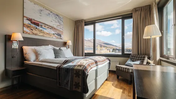 Ljust hotellrum med dubbelsäng, fåtölj och stora fönster som släpper in dagsljus och visar ett fjällandskap.