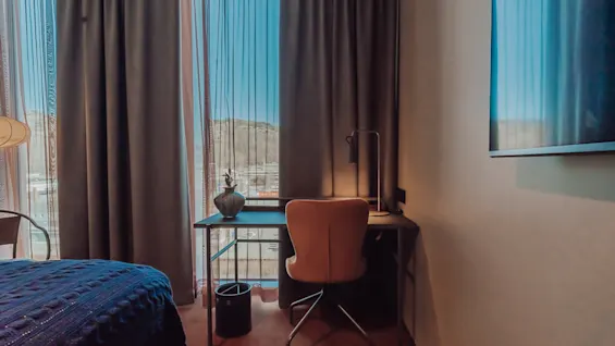 En skrivbord med en vas och lampa står mot ett fönster med gardiner, bredvid en säng med en blå filt. En stol står framför skrivbordet.