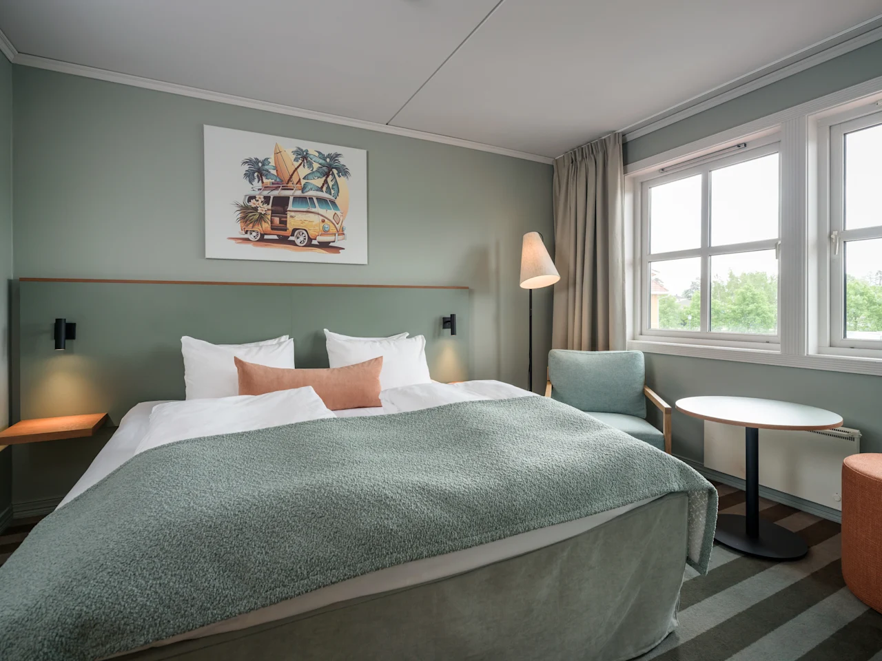 En pent senger med grønt sengeteppe står i sentrum av et moderne hotellrom, flankert av små nattbord. En gulvlampe står i nærheten, og et vindu viser grønt utenfor.