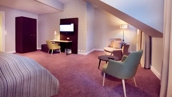 Et ryddig moderne hotellrom med mønstret vegg, en seng, stoler, et skrivebord, et klesskap og lamper, som viser komfortable møbler i et koselig, varm belyst rom.