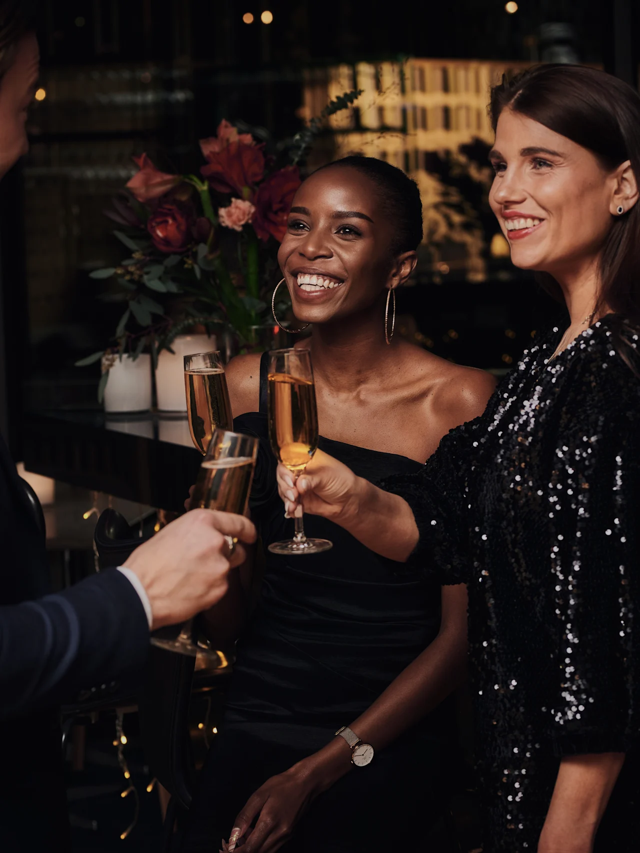 Tre personer i formella kläder skålar med champagneglas, ler varmt i en svagt upplyst, elegant lokal dekorerad med blommor och mjuk belysning.