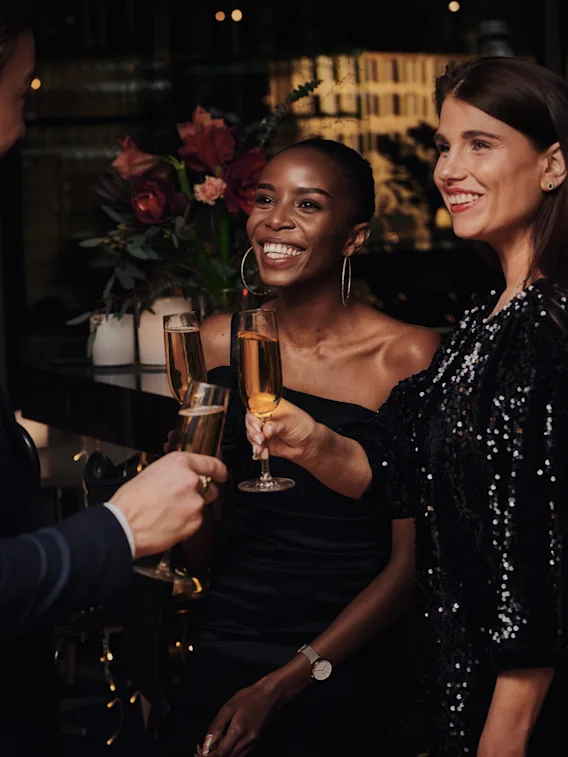 Three people in formal attire toast with champagne glasses, smiling warmly in a dimly lit, elegant venue decorated with flowers and soft lighting.