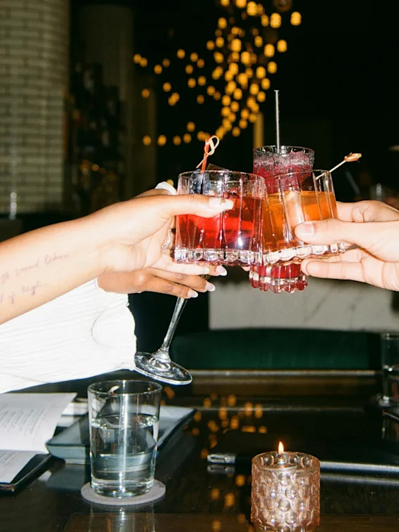 Glass fylt med fargerike cocktails klinker sammen i en feirende skål, omgitt av en svakt opplyst restaurant med hengende lys i bakgrunnen.