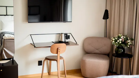 Et indbydende hotelværelse med et tv, et lille skrivebord til forfriskninger og en behagelig lænestol ved siden af en vase med friske blomster.