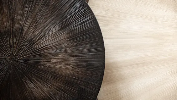Two round tables with radiating patterns, one dark brown and the other light beige, overlap slightly. They rest on a textured surface, creating a contrast in colors and textures.