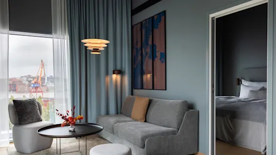 En grå soffa står i ett modernt vardagsrum med ett runt bord, golvlampa och abstrakt väggkonst. Stort fönster visar en stadsbild, mjukt ljus och en säng i angränsande rum.