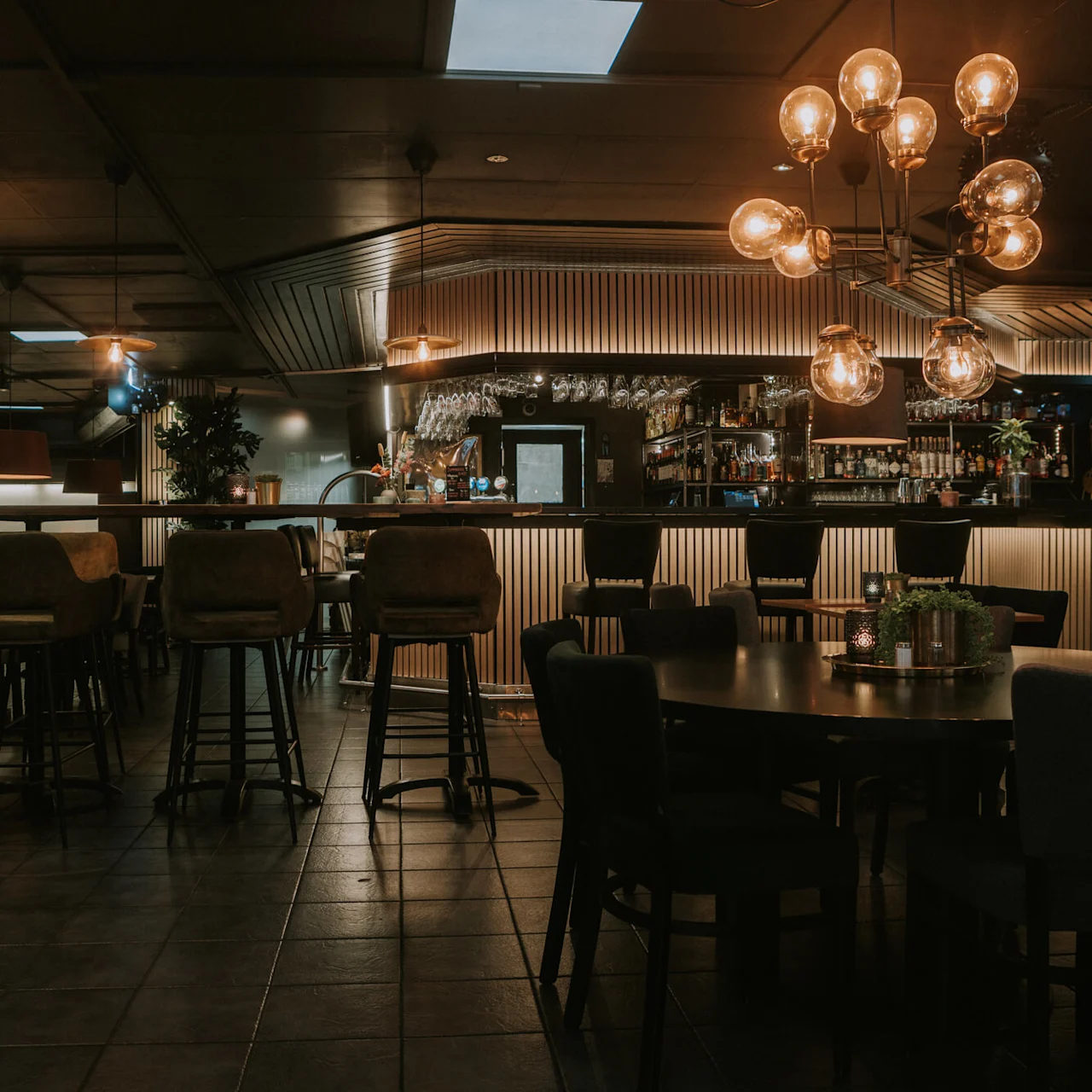 En dunkelt oplyst bar med stole, borde og pendellamper; ingen kunder, rolig atmosfære.