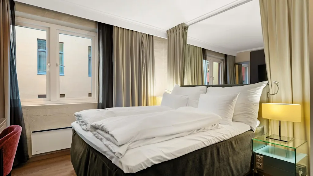 En pent oppredd seng med hvite sengetepper er i sentrum av et koselig rom, omgitt av gardiner, et vindu og et nattbord med lampe.