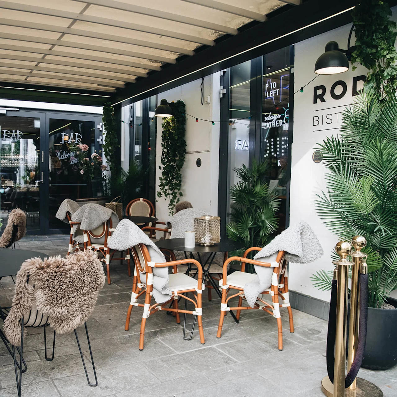 Utendørs terrasse med bord og stoler med skinn. Potteplanter gir grønt rundt. Tekst inkluderer: "BAR", "Welcome", "S TAY", "PER", "TO LEFT", "RODENS BISTRO".