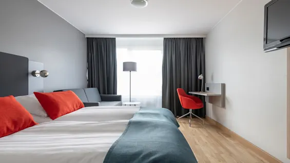 Ett hotellrum har en prydligt bäddad säng med orange kuddar, en grå soffa, ett skrivbord med en röd stol och en platt-TV på väggen, belyst av mjukt naturligt ljus.