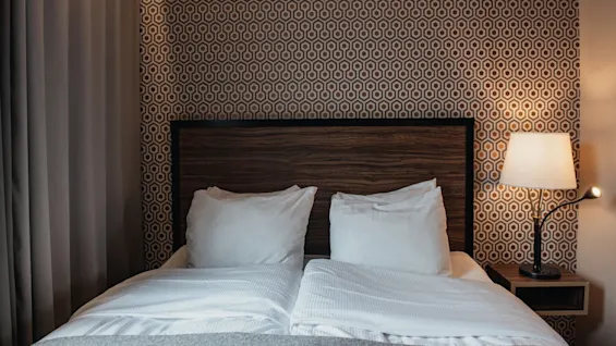Viihtyisä hotellihuone, jossa siististi pedattu sänky, puinen sängynpääty, kuviollinen tapetti ja lämmin yöpöytälamppu.