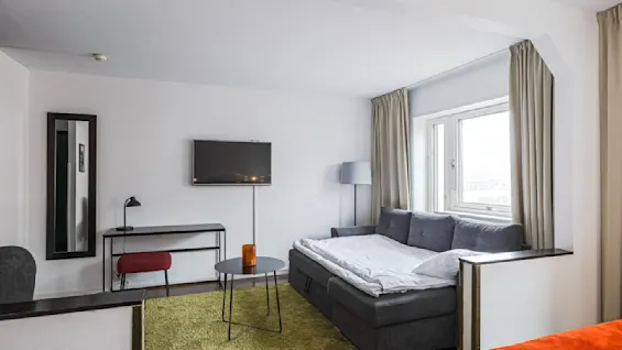 Et hyggelig moderne rom med en myk sovesofa ved et vindu, omgitt av beige gardiner. Et elegant bord holder et lys, som gir varme. En TV er montert på veggen over et minimalistisk skrivebord og en rød stol.