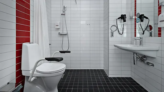 Toalett med armstöd står bredvid en dusch i ett vitt kaklat badrum. Ett draperi separerar duschutrymmet. En spegel och en hårtork är ovanför tvättstället, med röda accentplattor.