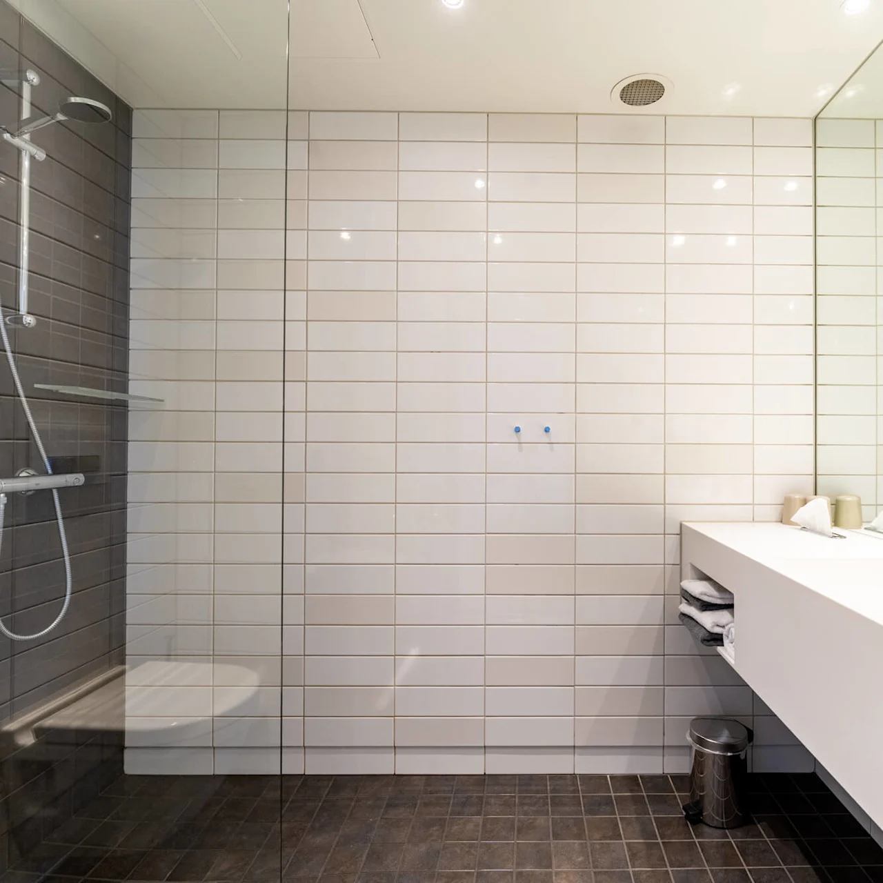 Et moderne badeværelse med en vægmonteret bruser til venstre, en hvid flisevæg som baggrund og en glat bordplade med dobbelte håndvaske til højre, udstyret med vandhaner og toiletartikler.
