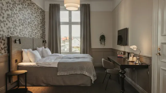 Ett mysigt hotellrum med en bekväm säng, ett skrivbord med TV och ett stort fönster med utsikt över en stad.