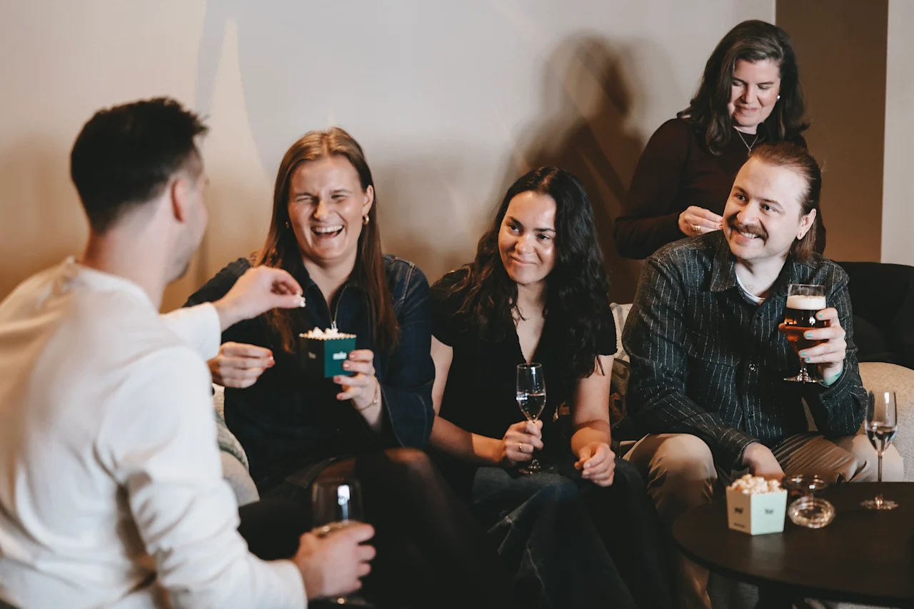 A group of five people sits and stands while smiling and chatting in a cozy room. Some hold drinks and small boxes, suggesting a relaxed social gathering.