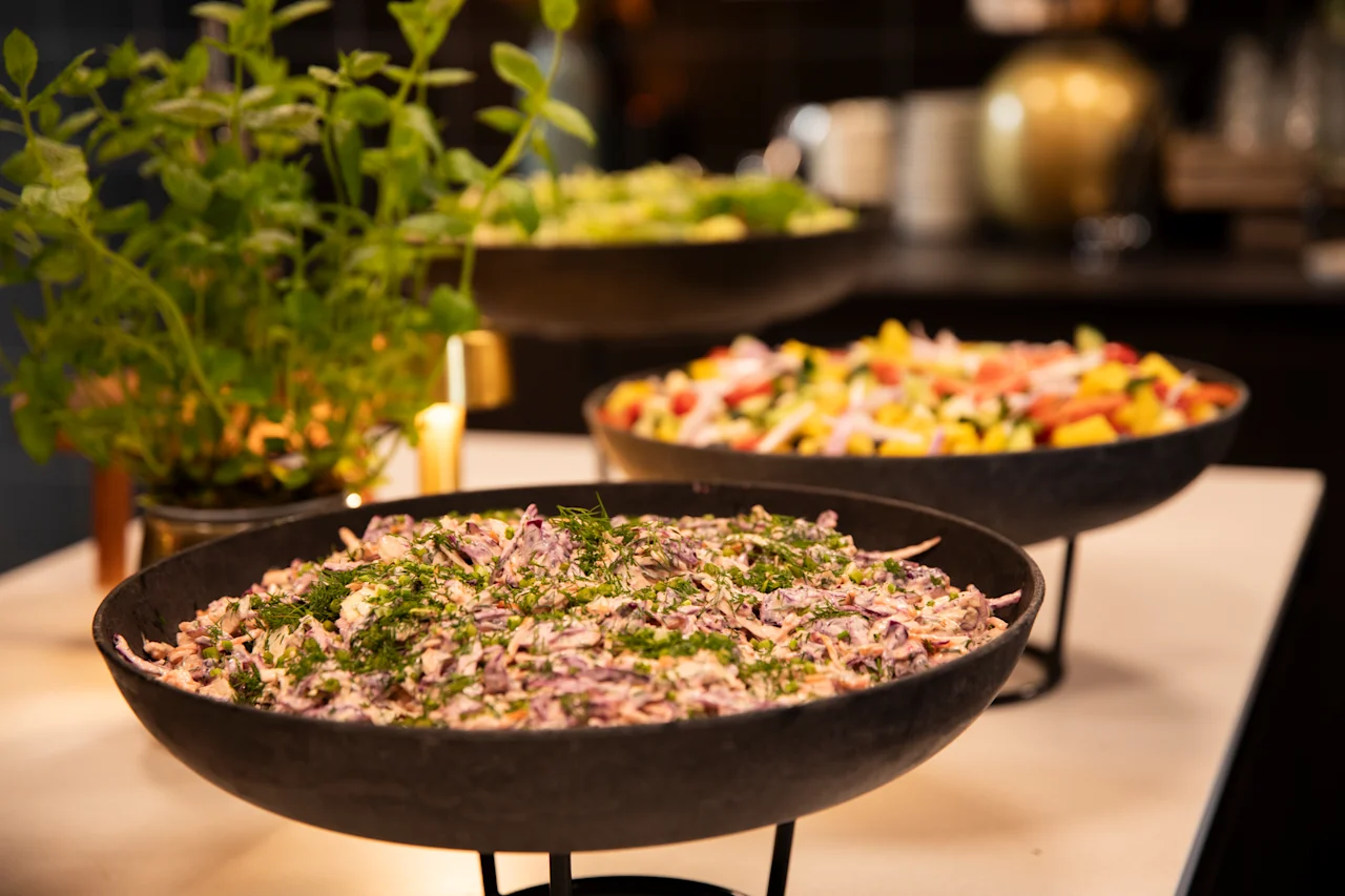 En buffé med tre skåler friske salater, inkludert coleslaw og en fargerik frukt- og grønnsakssalat, med en potteplante i bakgrunnen.