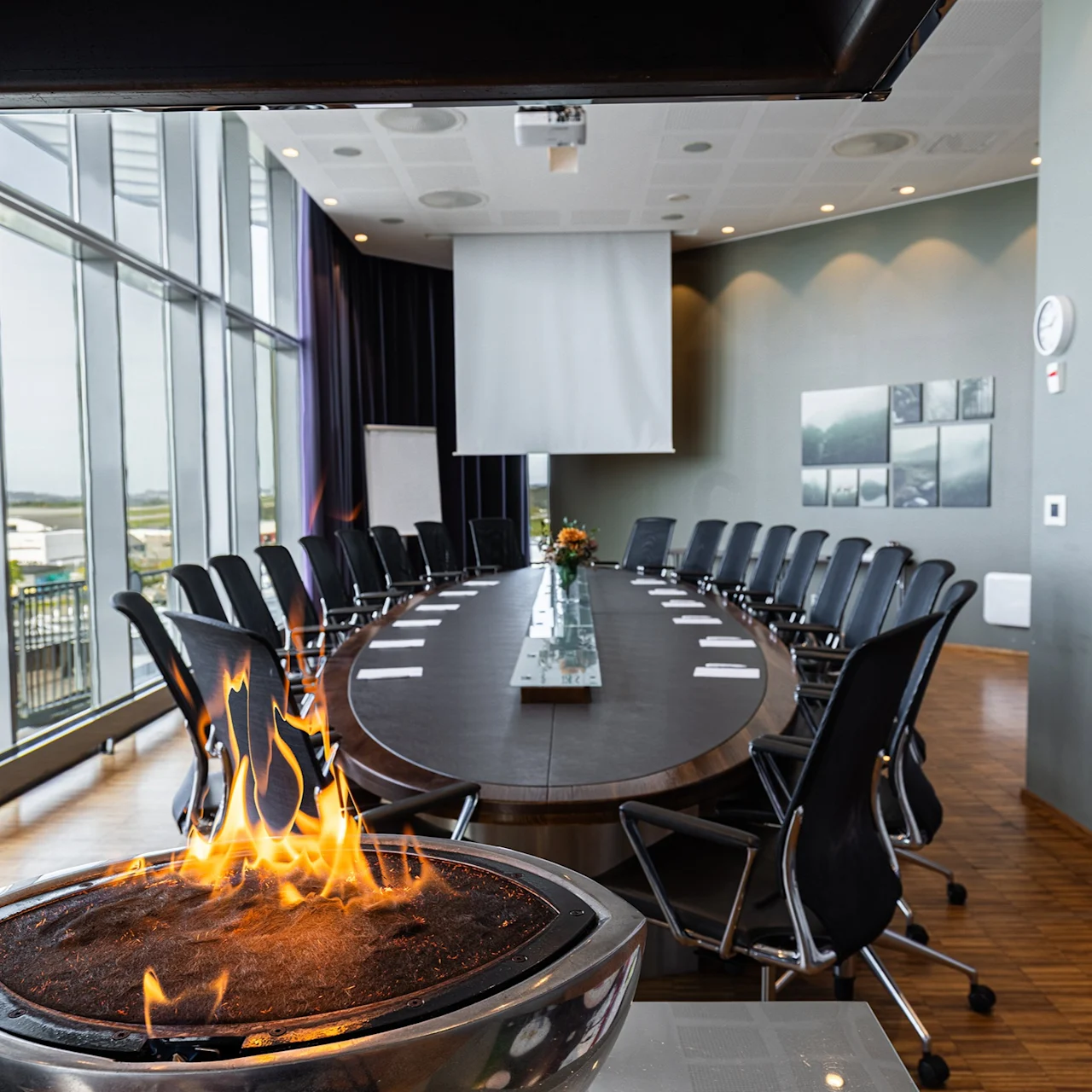 Fireplace crackling with flames, set in a modern conference room. Oval table surrounded by chairs, large windows overlook an outdoor view, with a white screen and artwork on the walls.