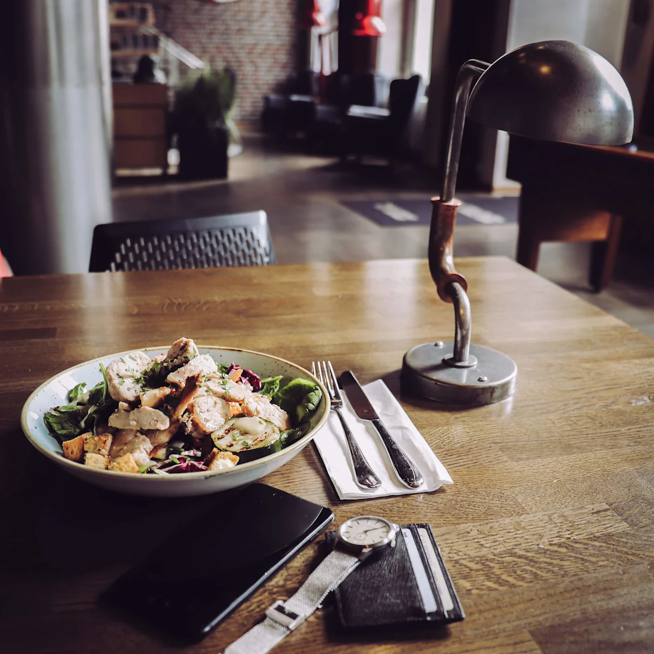 A salad bowl with chicken sits on a wooden table. A fork, knife, napkin, lamp, smartphone, wristwatch, and wallet are nearby. Background shows a dimly lit interior space.