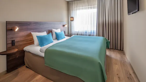 A neatly made bed with a wooden headboard and teal bedding is situated in a simple, modern room with light wood flooring, curtains, and a wall-mounted TV. 