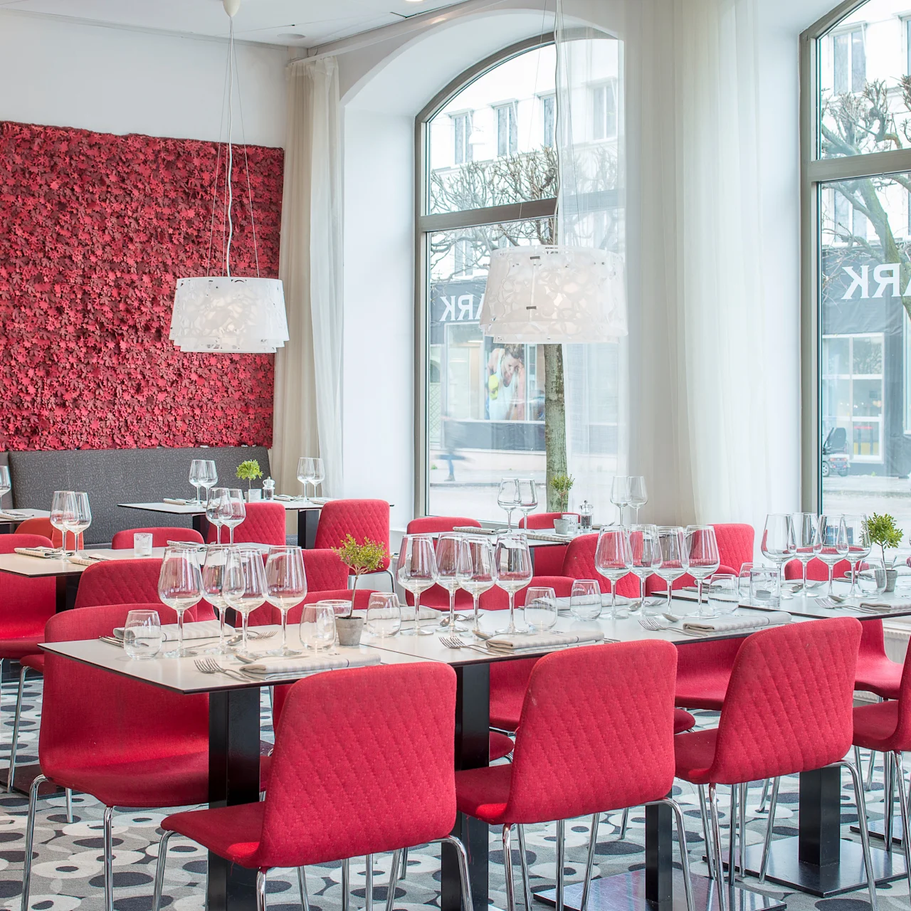 Elegante spisebord og røde stoler er dekket med glass i en moderne restaurant. Store vinduer gir utsikt over gaten utenfor. En teksturert rød vegg tilfører dekor.