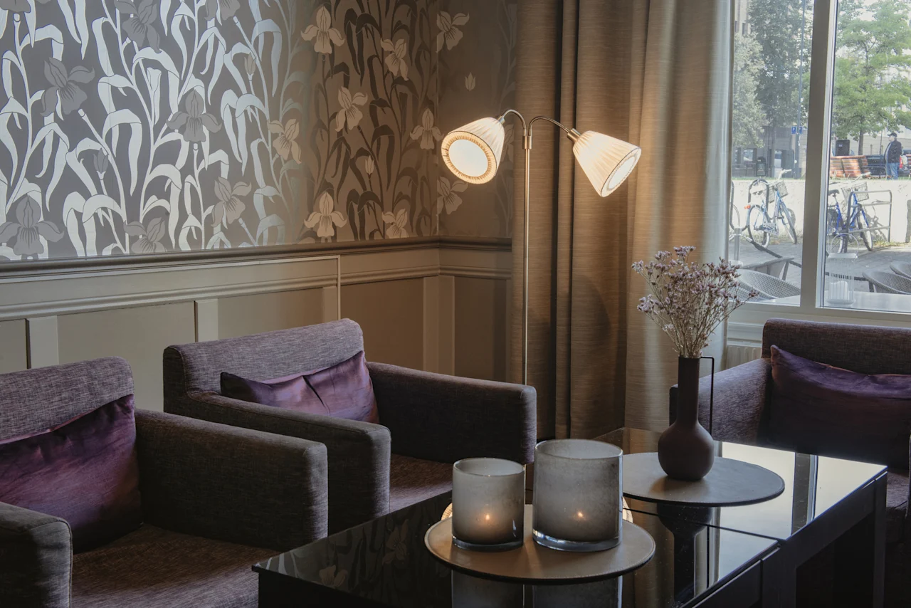 En bekväm hotellounge med fåtöljer, ett soffbord och en golvlampa. Blommig tapet och ett fönster mot en gata.